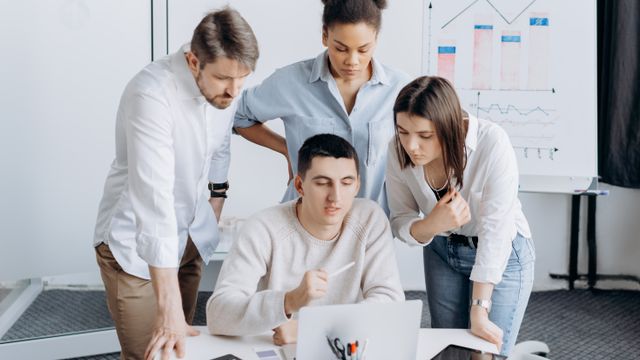 Three colleagues leaning in to look at a laptop with a fourth person at a desk.